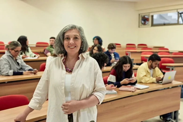 Žena stojí v univerzitní učebně s studenty na pozadí.