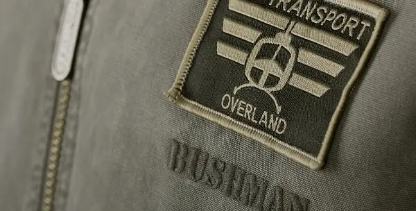 Detail šedého bundy s nášivkou Overland na zipu.