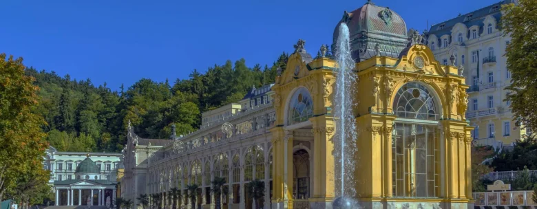 Žlutá lázeňská budova s fontánou na pozadí zelených hor.