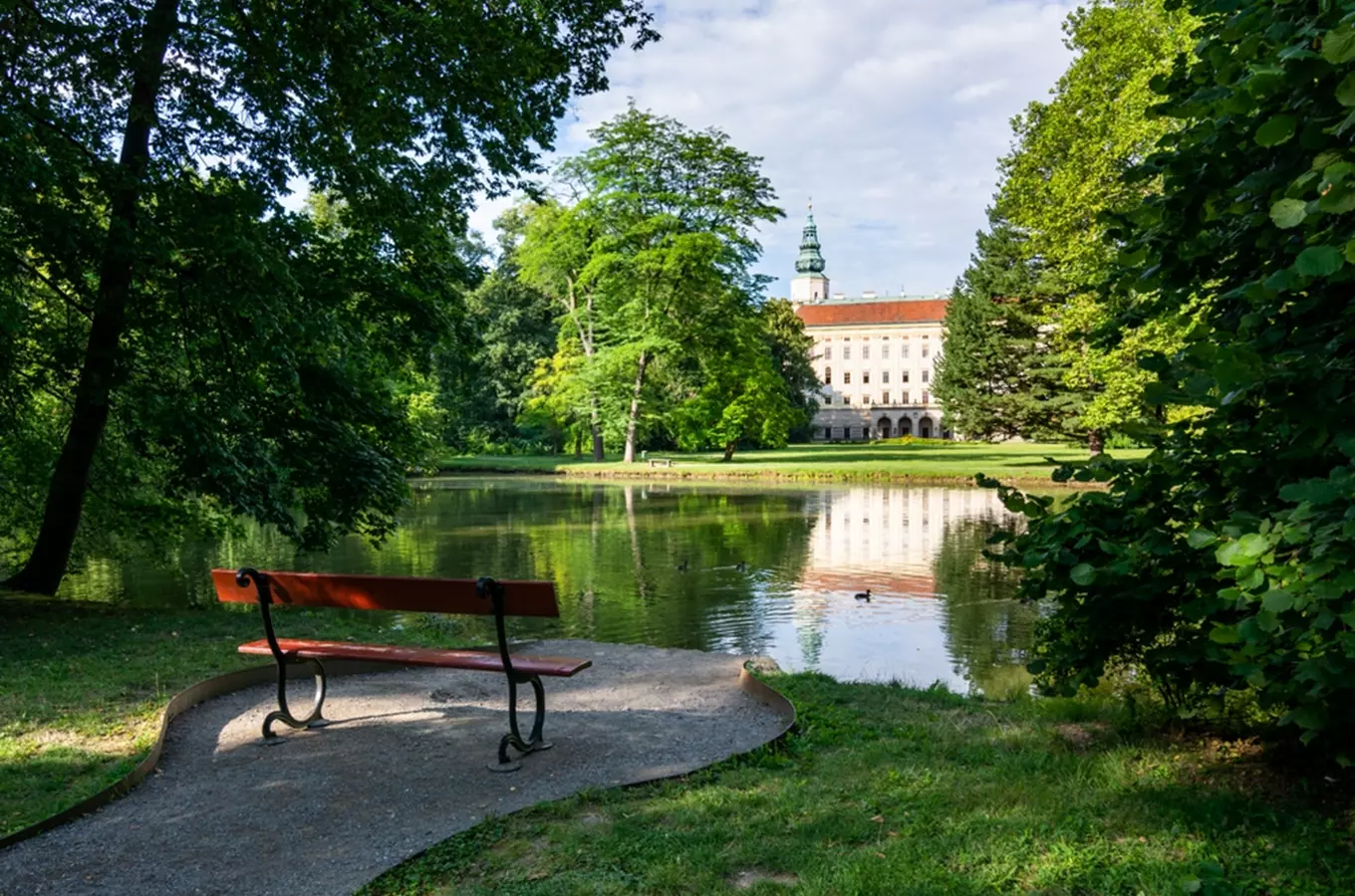 Pohled na zámek v Kroměříži s jezerem a lavičkou.