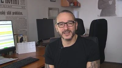 Muž v brýlích sedí za psacím stolem s počítačem.