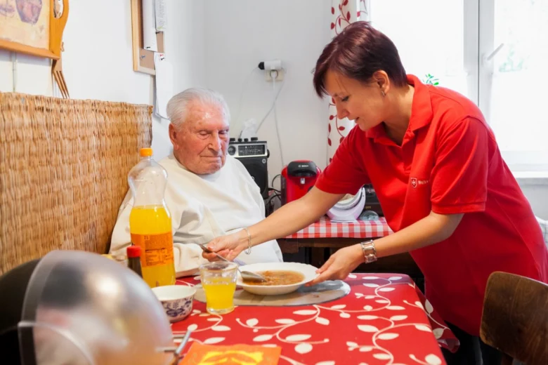 Péče o seniory: pracovnice pomáhá staršímu muži při jídle.