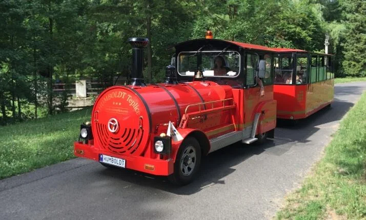 Červený turistický vlak s cestujícími na zalesněné trati.