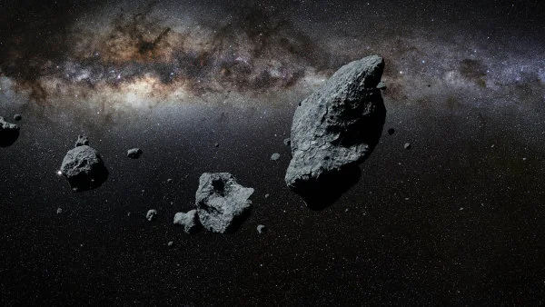 Skupina asteroidů plující ve vesmíru s Mléčnou dráhou na pozadí.