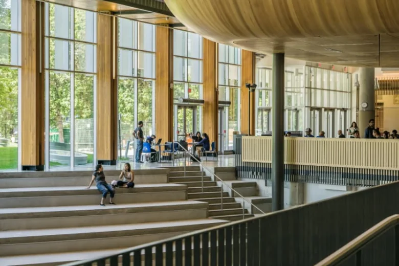 Moderní interiér univerzity s studenty na schodech a velkými okny.