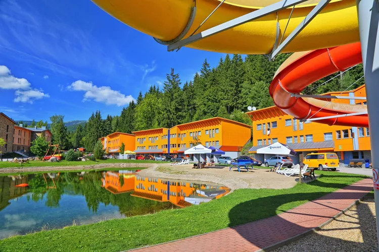 Aquapark v Česku s vodními skluzavkami a oranžovými budovami u jezírka.