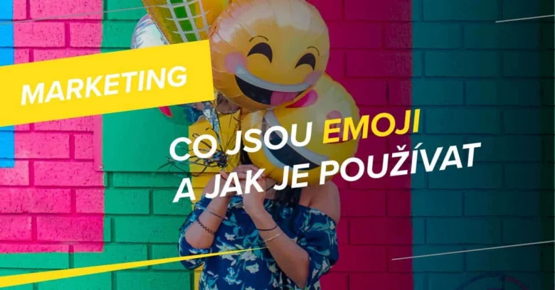 Osoba drží velké žluté balónky s tváří smajlíka před barevnou zdí.