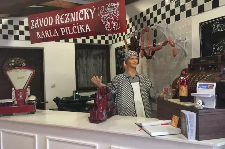 Manekýna v řeznictví s klobásami na háčcích pod červenou tabulí.