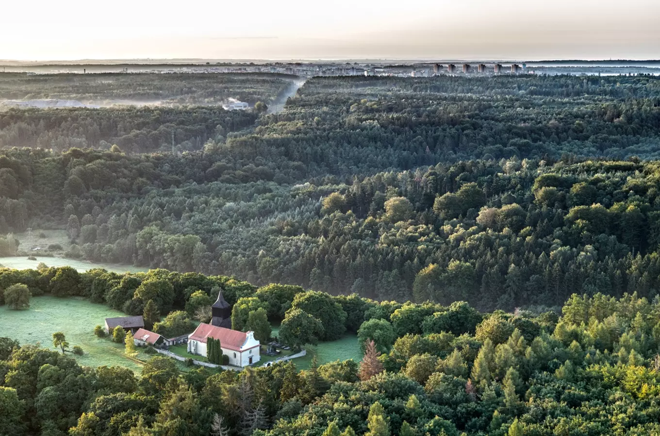 Pohled na les s kostelem v popředí a městem ve vzdálenosti.