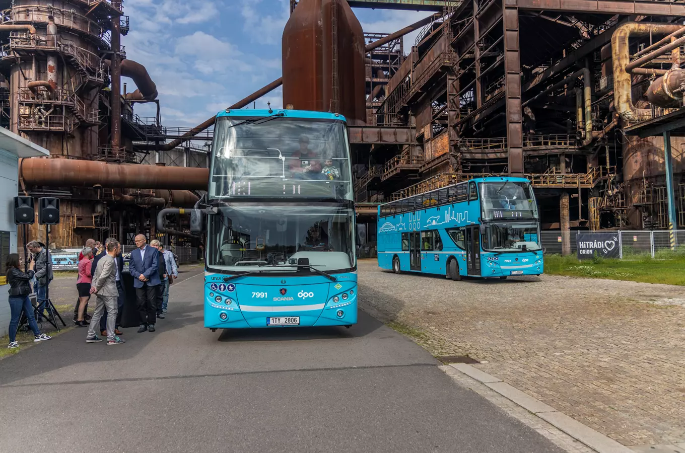 Dva modré dvoupodlažní autobusy před opuštěnou továrnou v Ostravě.