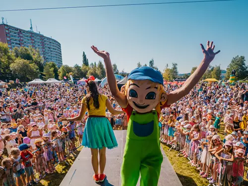 Maskot v zelených kalhotách mává davu dětí na venkovním slavnostním programu.