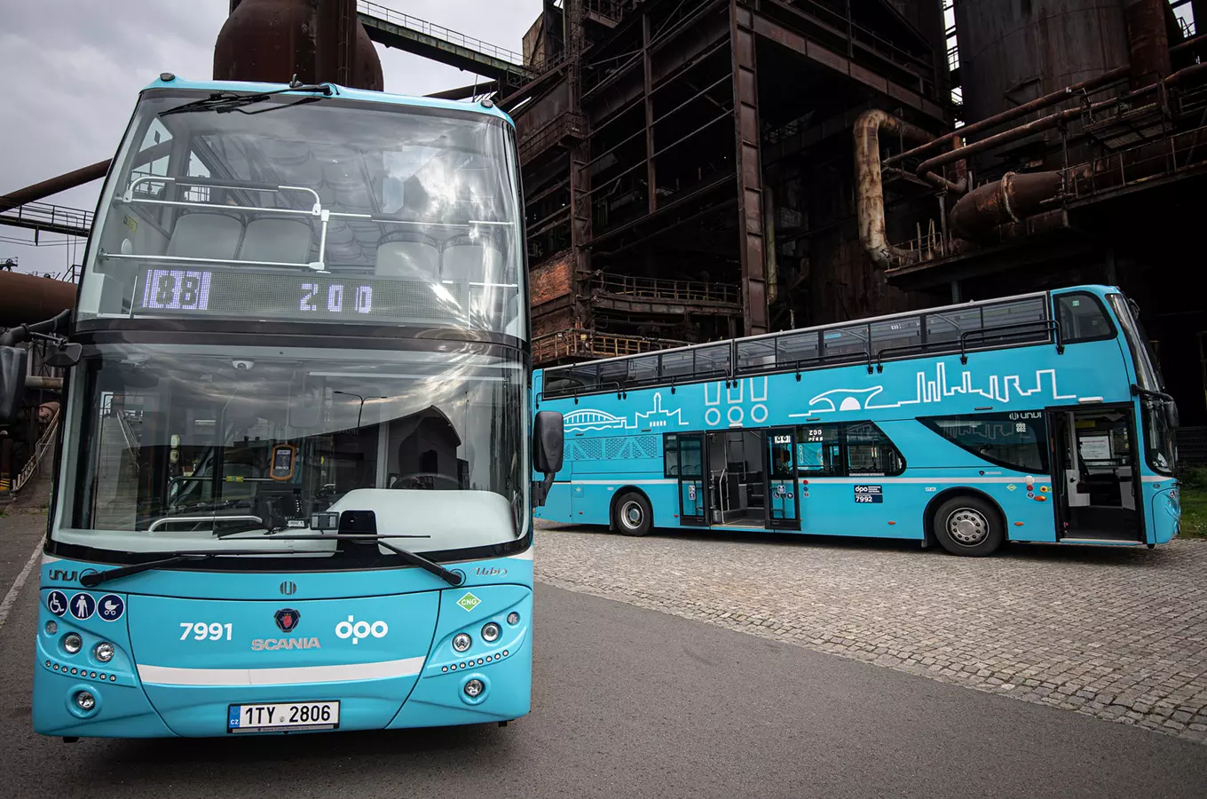 Dva modré dvoupodlažní autobusy před průmyslovou budovou v Ostravě.