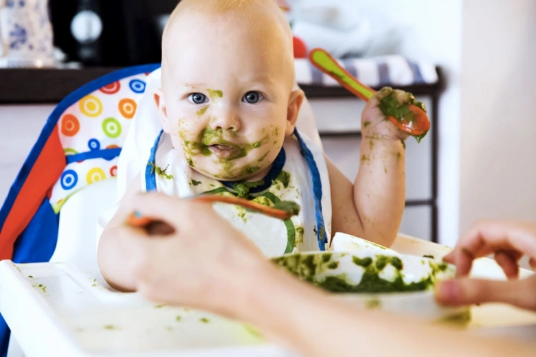 Babička krmit zeleninovou kaší dítě v jídelní židli.