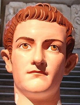 Socha císaře Kaligyuly s červenými vlasy na historickém pozadí.