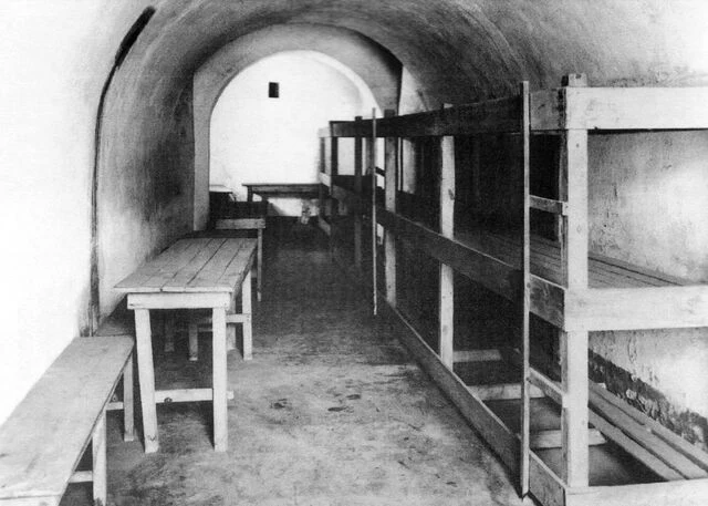 Interiér věznice s dřevěnými postelemi v architektonickém tunelu.