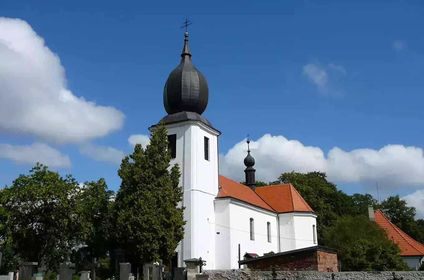 Bílá kostel s cibulovou kupolí na hřbitově pod modrou oblohou.