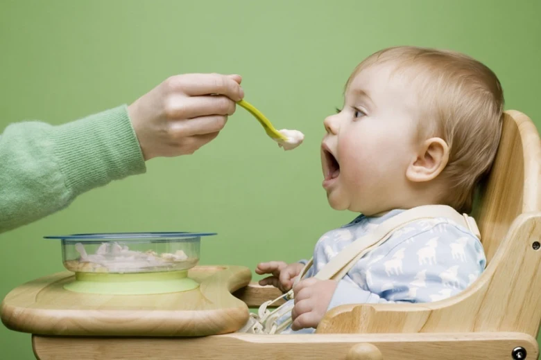 Dítě v jídelní židli přijímá příkrmy lžičkou.