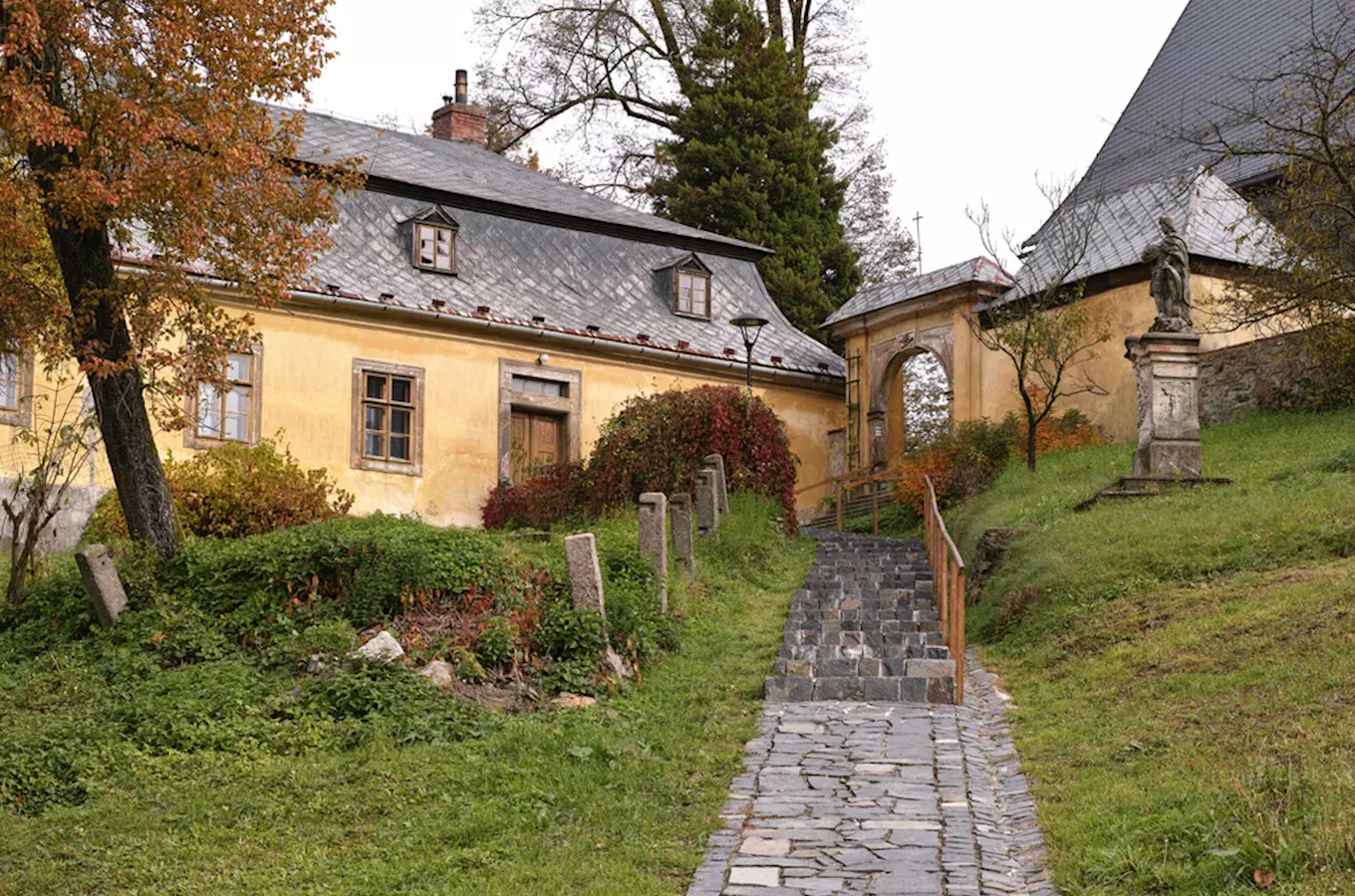 Historická žlutá budova s kamennou cestou sochou v podzimním prostředí.