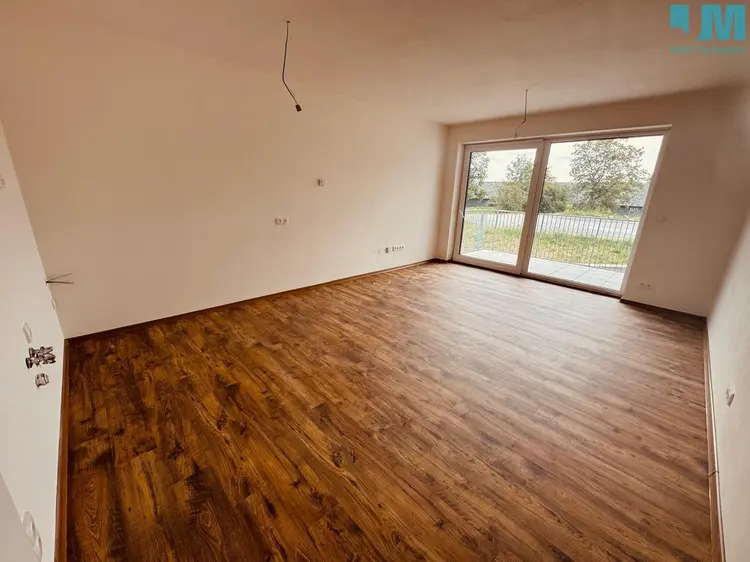 Prázdný byt s dřevěnou podlahou a výhledem na zahradu přes balkon.
