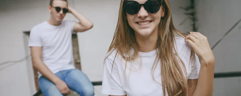 Usměvavá žena v slunečních brýlích s mužem na pozadí.