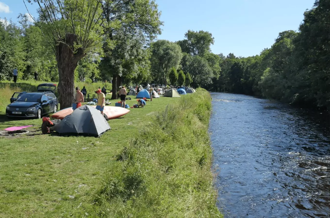 Kempování na břehu řeky s stany a lidmi v přírodě.