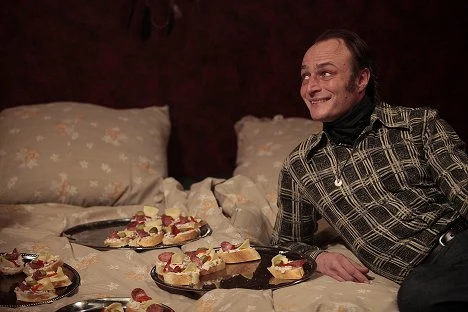 Herec Karel Dobrý sedí na posteli s talíři jídla.
