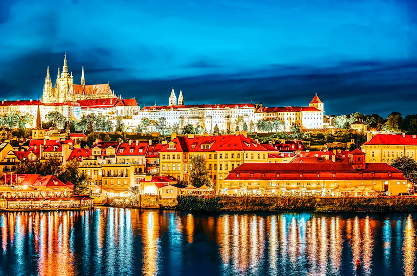 Pražský hrad osvětlený v noci s odrazem ve vodě řeky.