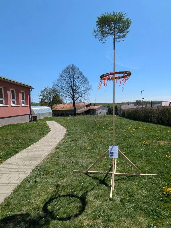 Tradiční májka s věncem a stromovou korunou na venkovském pozadí.