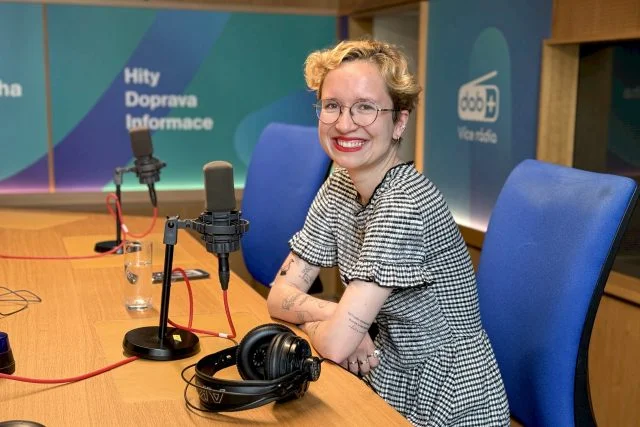 Žena sedí v rozhlasové studii s mikrofony a sluchátky.