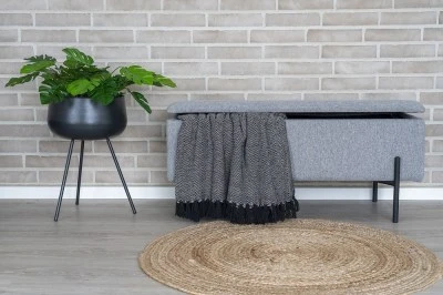 Šedá úložná lavice s dekou v předsíni vedle rostliny.
