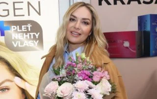 Anna Polívková s květinami na akci vedle reklamních plakátů.