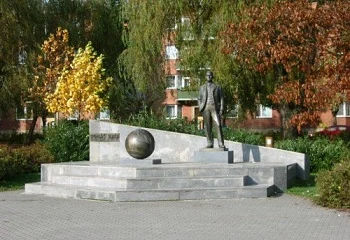 Bronzová socha muže s kulí na kamenném podstavci v parku.