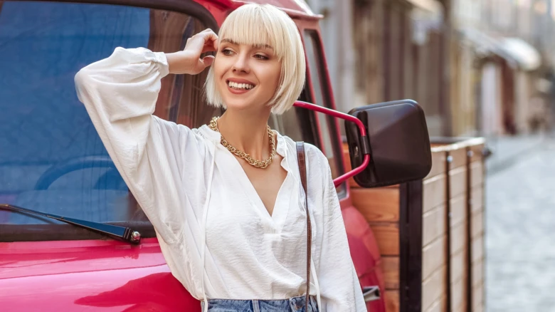 Žena s blond bob účesem stojí vedle červeného auta.