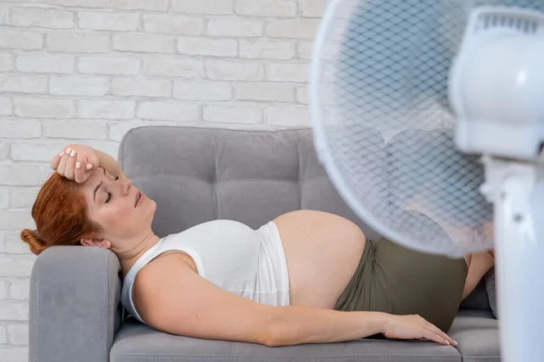 Těhotná žena leží na gauči s ventilátorem kvůli přehřívání.