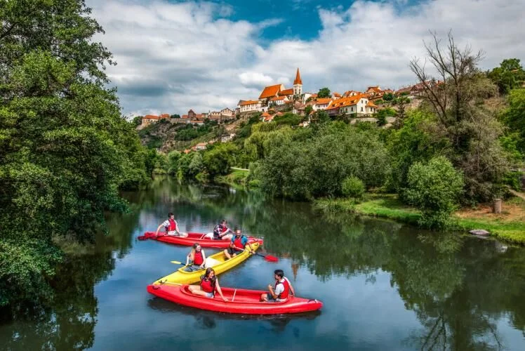 Skupina lidí v kajakech pluje řekou směrem k historickému městečku na kopci.