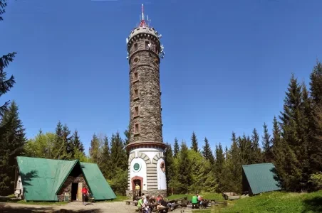 Kamenná rozhledna v lesu s turistickými chatami pod modrou oblohou.