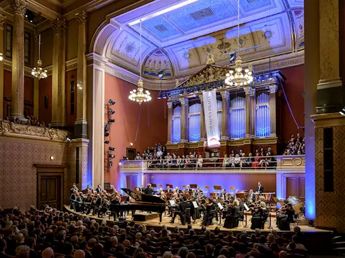 Orchester hraje v pražské koncertní síni před plným divákem.