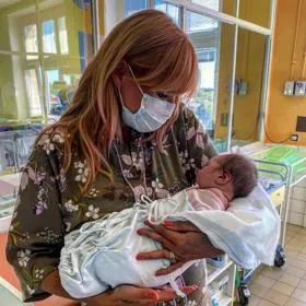 Žena s rouškou drží novorozence v nemocničním pokoji.