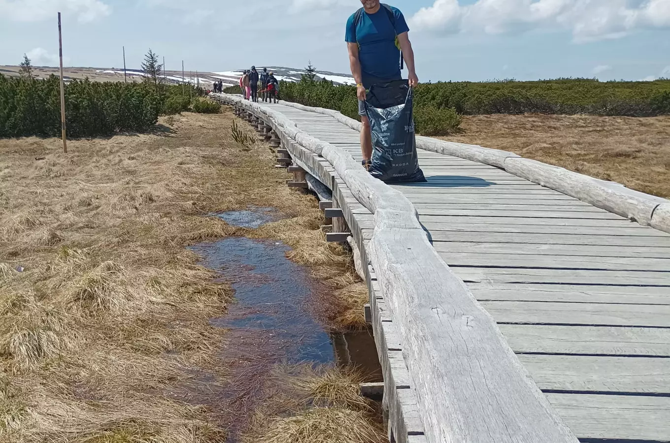 Turista s pytlem na odpadky kráčí po dřevěné chodníku v Peci pod Sněžkou.