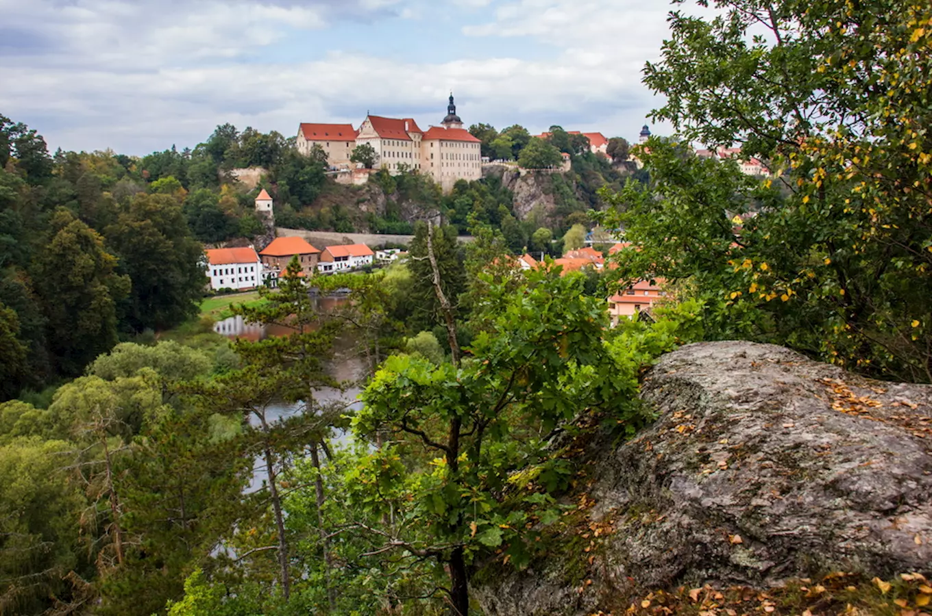 Zámek v Bečyni nad řekou s okolními domy a lesy.